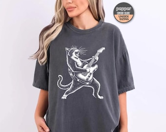 May include: Dark gray t-shirt featuring a white illustration of a cat playing an electric guitar. The cat is in a dynamic pose, with its mouth open as if singing. The shirt has a relaxed fit. The text "pepper" is visible on a circular tag.