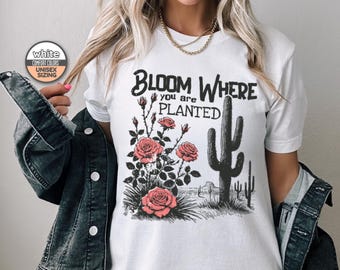 May include: White t-shirt with a graphic print of a cactus and roses. The text "Bloom Where You Are Planted" is printed on the shirt.