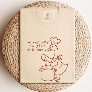 May include: Cream-colored t-shirt with a whimsical illustration of a goose wearing a chef's hat and stirring a pot. The text "on my way to stir THE POT" is printed above the goose. The shirt has a crew neck and a tag at the neckline.