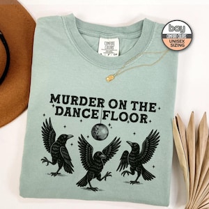 May include: A light green unisex sweatshirt with the phrase "MURDER ON THE DANCE FLOOR" in black text. The design features three black crows, a disco ball, and stars. A gold necklace is draped on the shirt.