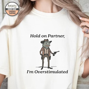 May include: A white t-shirt with the text "Hold on Partner, I'm Overstimulated" and a cartoon image of a frog wearing a cowboy hat and holding a gun.
