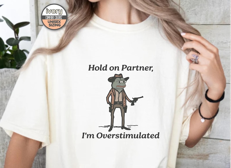 May include: A white t-shirt with the text "Hold on Partner, I'm Overstimulated" and a cartoon image of a frog wearing a cowboy hat and holding a gun.
