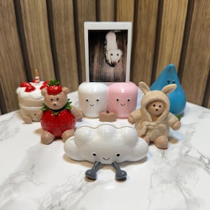 May include: A collection of small decorative figurines including a strawberry-themed bear, a white cloud with grey legs, a beige bear in a hooded outfit, a blue teardrop shape, a cake, and two marshmallow-like figures, all surrounding a small photo.
