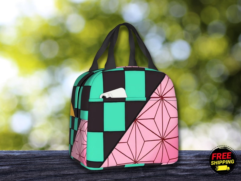 K&ouml;nnte beinhalten: Eine schwarz-gr&uuml;ne karierte Lunch-Tasche mit einem pink-wei&szlig;en geometrischen Muster an der Seite. Die Tasche hat einen schwarzen Griff und eine kleine Tasche auf der Vorderseite.