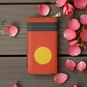 Puede incluir: Una cartera roja con un círculo amarillo y un diseño de rayas negras. La cartera tiene una cremallera dorada y es de forma rectangular. Pétalos de flores rosas están esparcidos alrededor de la cartera sobre una superficie de madera.