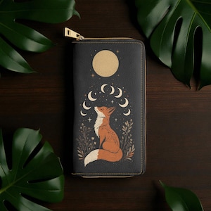 Könnte beinhalten: Schwarze Geldbörse mit goldenem Reißverschluss, mit einer Illustration eines Fuchses unter einem Vollmond und Halbmonden. Der Fuchs ist orange und weiß, umgeben von Pflanzen.