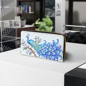 May include: A rectangular wallet with a white background featuring a detailed illustration of a peacock perched on a branch with light purple flowers. The peacock has vibrant blue and green feathers with distinct eye patterns. It has a black zip.