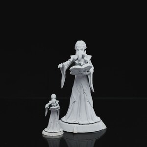 May include: Two white painted figurines of a female wizard, one larger than the other, both holding a book and standing on a base.