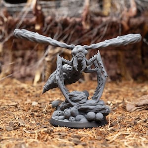 May include: A gray 3D printed miniature of a creature with wings, six legs, and a segmented body. The creature is posed in a flying position with its wings spread wide. The miniature is set on a base with a rocky terrain and mushrooms.