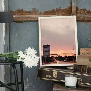 May include: A framed photograph of a city skyline at sunset. The photo shows a tall building, a bridge, and a body of water. The photo is hanging on a wall with a wooden box and a candle below it.
