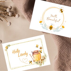 Puede incluir: Dos tarjetas de felicitaci&oacute;n con marcos dorados y dise&ntilde;os con tem&aacute;tica de abejas. La primera tarjeta dice "Hello Beautiful" con una abeja y flores. La segunda tarjeta dice "Hello Lovely" con una abeja, un tarro de miel y flores.