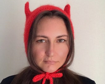 May include: A red knitted hat with horns, resembling a devil's hat, is worn by a person with long brown hair. The hat has a red knitted bow tied at the neck.