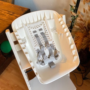 Op de afbeelding: Een witte kinderstoel met een grijze veiligheidsgordel. De stoel heeft een dienblad en een voetensteun. De stoel is gemaakt van plastic en is ontworpen voor baby's en peuters.