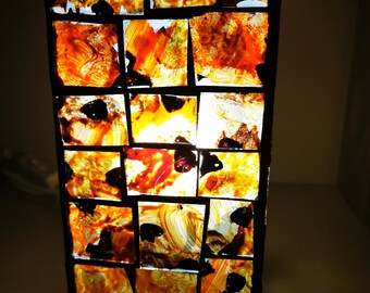 Pode incluir: Um abajur retangular feito de vidro colorido com um padrão de mosaico de quadrados marrons, laranja e amarelos. A luz brilha através do vidro, criando um brilho quente.