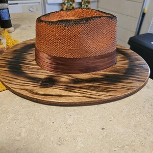 May include: A brown and black straw hat with a brown ribbon band, sitting on a wooden base with a burnt finish.