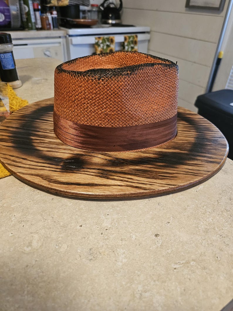 May include: A brown and black straw hat with a brown ribbon band, sitting on a wooden base with a burnt finish.