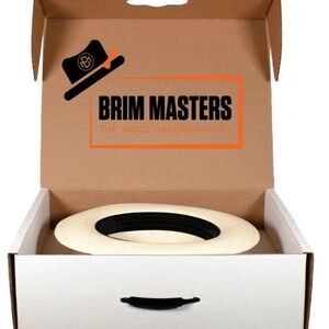 Puede incluir: Una caja de sombrero blanca con una manija negra. La caja est&aacute; abierta y un sombrero de paja blanco est&aacute; adentro. La caja tiene una tapa de cart&oacute;n marr&oacute;n con el texto "BRIM MASTERS THE WOOD HABERDASHERY" impreso en ella.