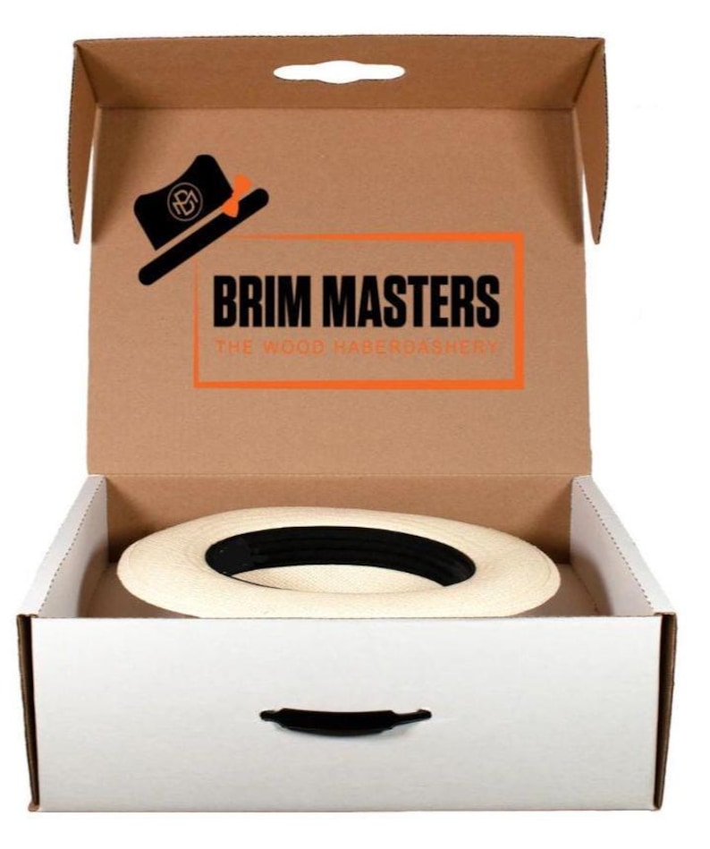 Puede incluir: Una caja de sombrero blanca con una manija negra. La caja est&aacute; abierta y un sombrero de paja blanco est&aacute; adentro. La caja tiene una tapa de cart&oacute;n marr&oacute;n con el texto "BRIM MASTERS THE WOOD HABERDASHERY" impreso en ella.