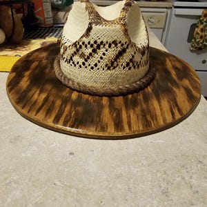 Puede incluir: Un sombrero de vaquero de paja blanca con una banda de cuero marr&oacute;n. El sombrero est&aacute; sentado en un soporte de madera con una mancha marr&oacute;n oscura.