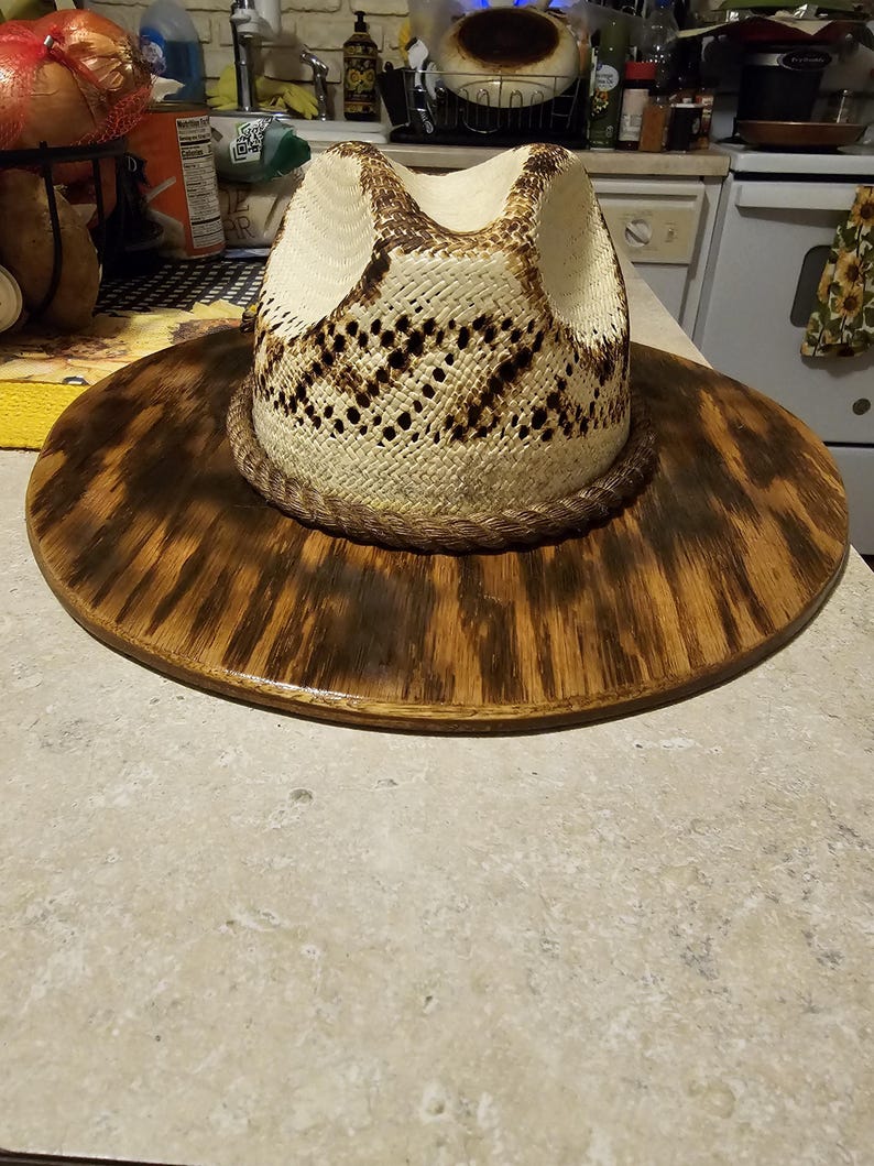 Puede incluir: Un sombrero de vaquero de paja blanca con una banda de cuero marr&oacute;n. El sombrero est&aacute; sentado en un soporte de madera con una mancha marr&oacute;n oscura.