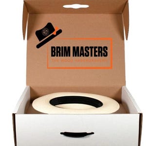 May include: A white hat with a black band is inside a cardboard box with a white interior. The box has a handle on the top and a logo that says "BRIM MASTERS THE WOOD HABERDASHERY".