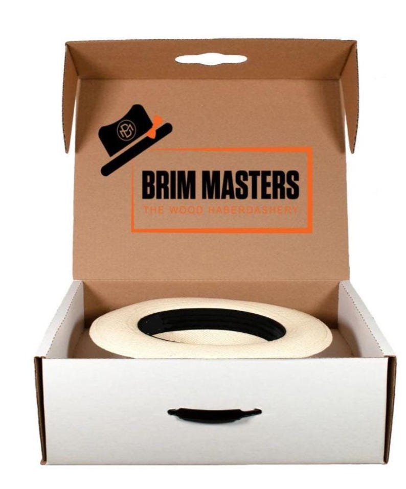 May include: A white hat with a black band is inside a cardboard box with a white interior. The box has a handle on the top and a logo that says "BRIM MASTERS THE WOOD HABERDASHERY".
