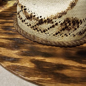 Puede incluir: Un sombrero de paja blanco con un dise&ntilde;o tejido marr&oacute;n sobre una base de madera marr&oacute;n oscuro.