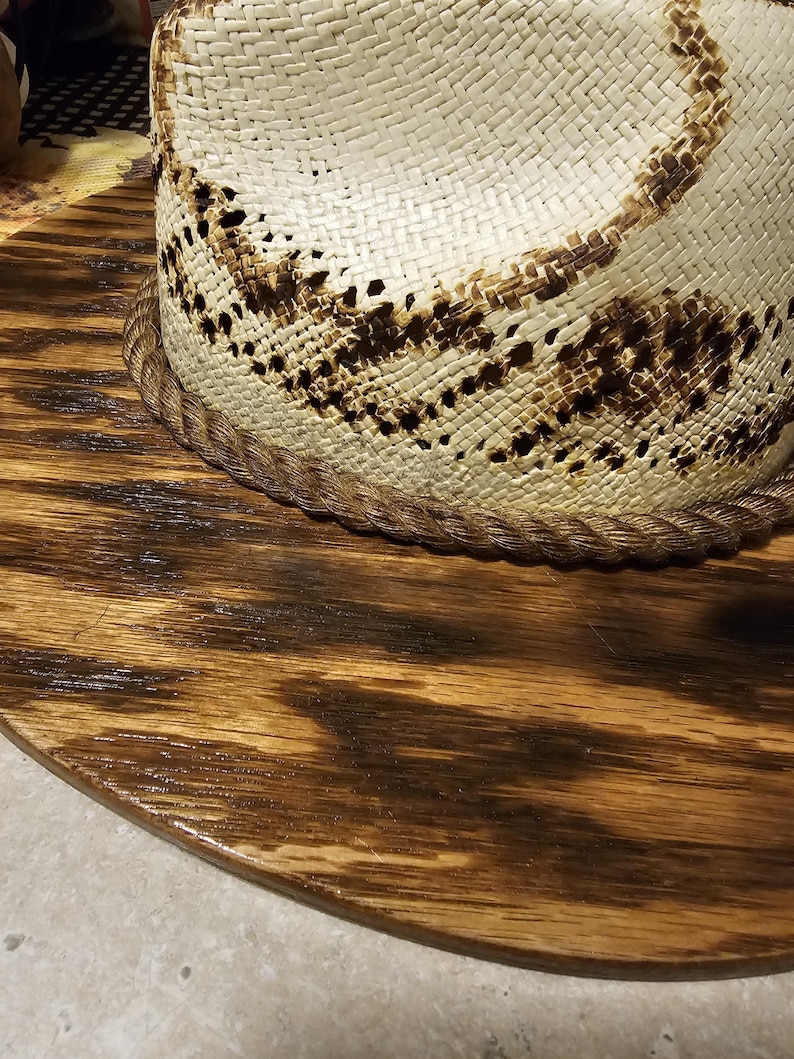 Puede incluir: Un sombrero de paja blanco con un dise&ntilde;o tejido marr&oacute;n sobre una base de madera marr&oacute;n oscuro.