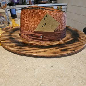 May include: A brown and orange straw hat with a brown ribbon and a card that says "A & 8" on it. The hat is sitting on a wooden surface.