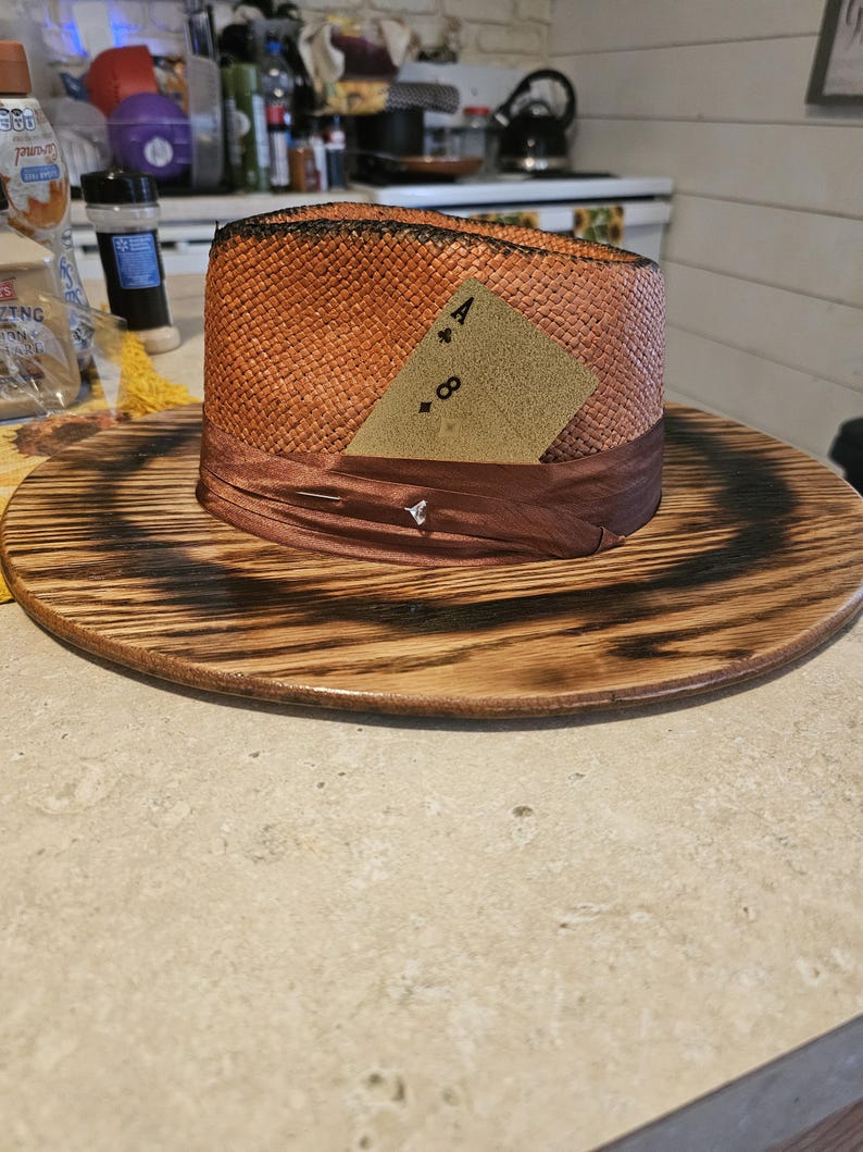 May include: A brown and orange straw hat with a brown ribbon and a card that says "A & 8" on it. The hat is sitting on a wooden surface.