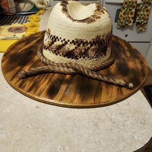 Puede incluir: Un sombrero de vaquero de paja blanca con una banda de cuerda marr&oacute;n y un dise&ntilde;o estampado marr&oacute;n y blanco. El sombrero se muestra en una base redonda de madera.