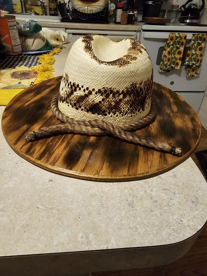 Puede incluir: Un sombrero de vaquero de paja blanca con una banda de cuerda marr&oacute;n y un dise&ntilde;o estampado marr&oacute;n y blanco. El sombrero se muestra en una base redonda de madera.