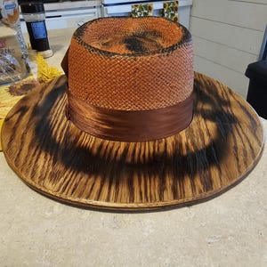 May include: A wide-brimmed hat with a brown woven crown and a brown ribbon. The hat has a wooden base with a dark brown finish.