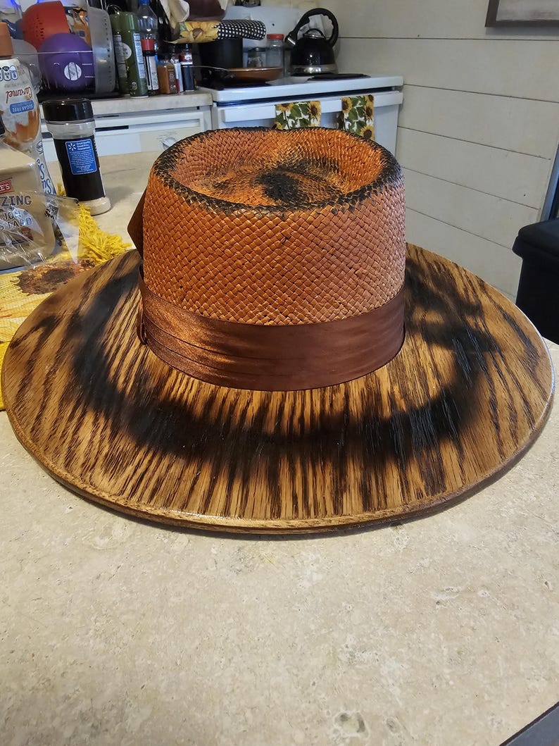 May include: A wide-brimmed hat with a brown woven crown and a brown ribbon. The hat has a wooden base with a dark brown finish.
