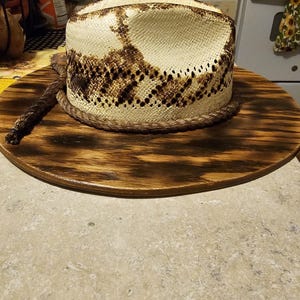 Puede incluir: Un sombrero de vaquero blanco con un patr&oacute;n de piel de serpiente marr&oacute;n en la corona. El sombrero est&aacute; sentado sobre una base de madera.