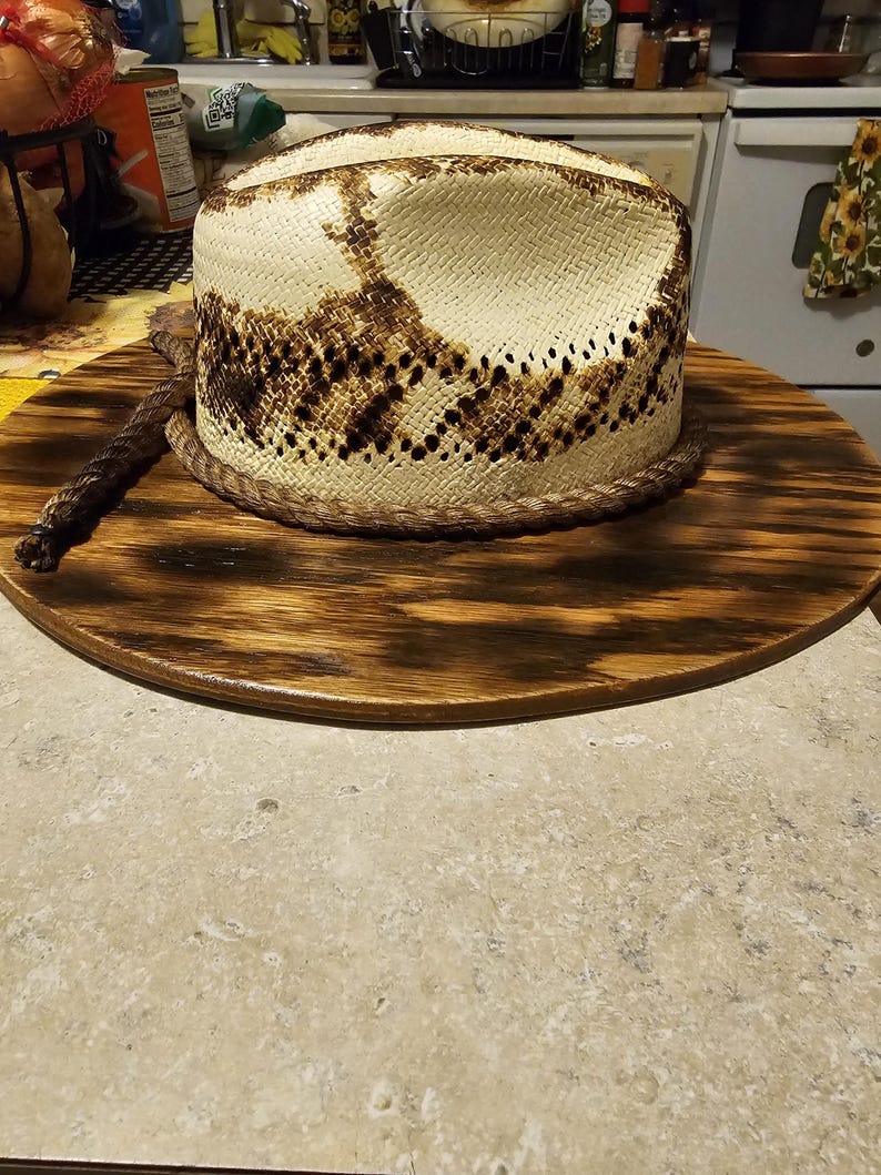 Puede incluir: Un sombrero de vaquero blanco con un patr&oacute;n de piel de serpiente marr&oacute;n en la corona. El sombrero est&aacute; sentado sobre una base de madera.