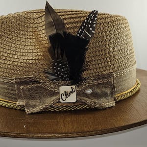 Puede incluir: Un sombrero fedora de paja marrón con una cinta marrón y un ribete dorado. El sombrero tiene un lazo marrón con una etiqueta blanca que dice "Clint". Una pluma negra con manchas blancas está unida al sombrero.