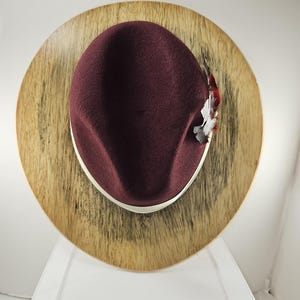 May include: A burgundy felt fedora hat with a white band and a small feather accent. The hat is displayed on a wooden hat stand.