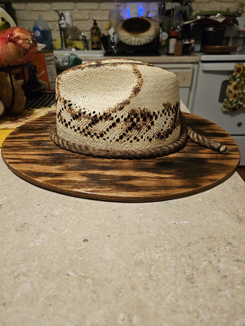 Puede incluir: Un sombrero de vaquero de paja blanca con una banda tejida marr&oacute;n y un patr&oacute;n marr&oacute;n y negro. El sombrero est&aacute; sentado sobre una base de madera.