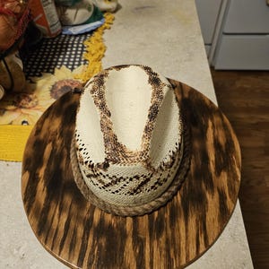Puede incluir: Un sombrero de vaquero de paja blanca con una banda de cuerda marr&oacute;n, exhibido en un perchero de sombrero de madera con un acabado de madera de grano marr&oacute;n oscuro.