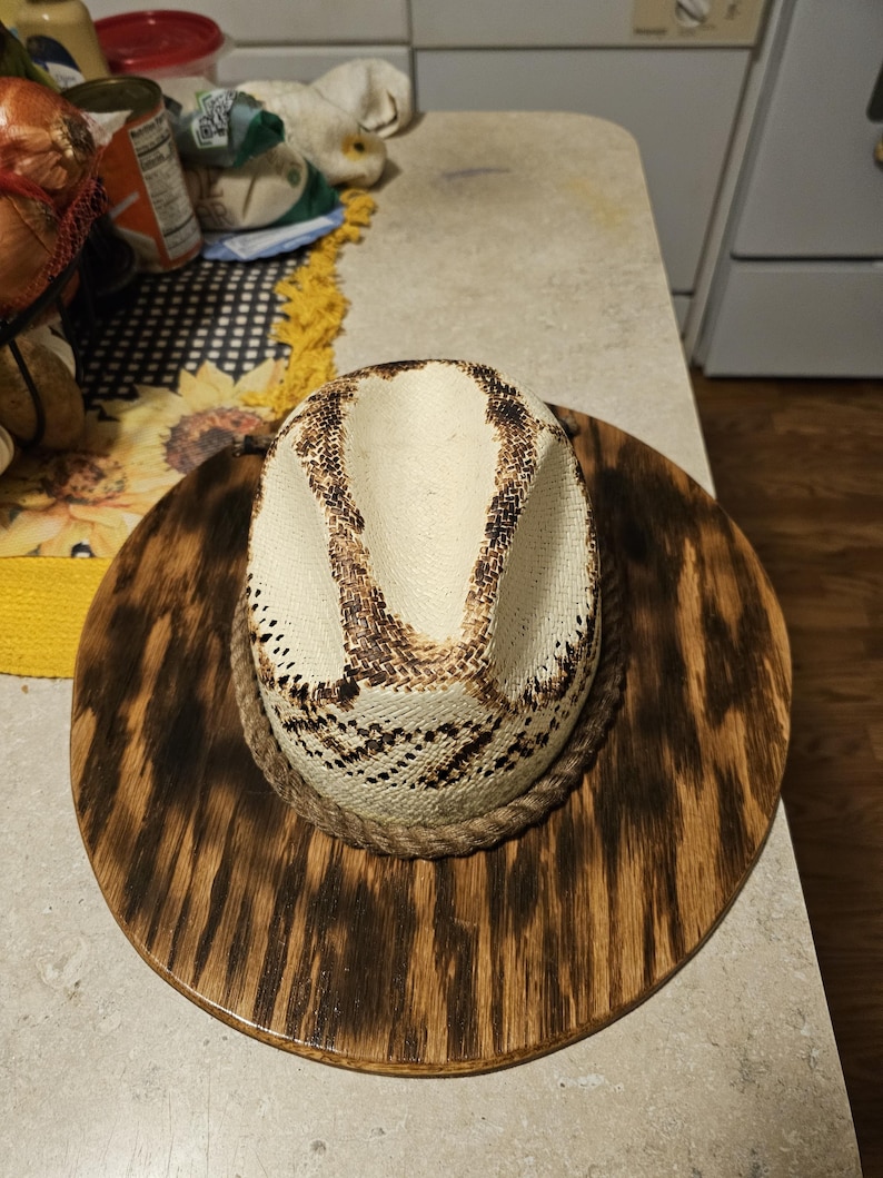 Puede incluir: Un sombrero de vaquero de paja blanca con una banda de cuerda marr&oacute;n, exhibido en un perchero de sombrero de madera con un acabado de madera de grano marr&oacute;n oscuro.