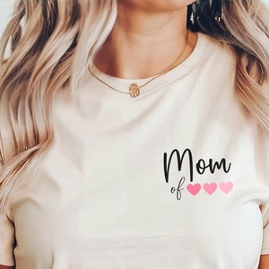 Könnte beinhalten: Ein beiges T-Shirt mit dem Text "Mom of" und drei rosa Herzen. Das Shirt wird von einer Person mit langen blonden Haaren getragen.