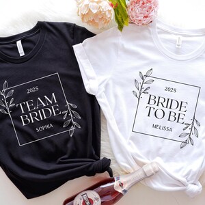 May include: Two white t-shirts with black text and floral designs. The text on one shirt says "2025 Team Bride Sophia" and the text on the other shirt says "2025 Bride to Be Melissa".