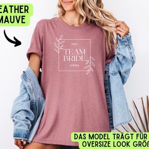 May include: Heather mauve t-shirt with a white graphic that says "2025 Team Bride Sophia". The shirt is being modelled by a person wearing a denim jacket.