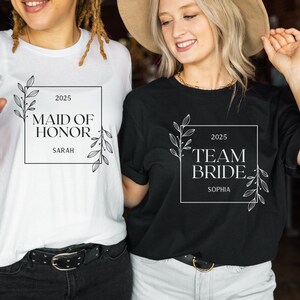 May include: Two white t-shirts with black text and floral designs. The first shirt says "Maid of Honour 2025 Sarah". The second shirt says "Team Bride 2025 Sophia".