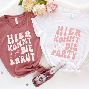 Könnte beinhalten: Zwei weiße T-Shirts mit rosa Schriftzug, auf denen "Hier kommt die Party" und "Hier kommt die Braut" steht. Die Shirts liegen auf einem weißen, flauschigen Untergrund mit rosa Konfetti und einer Flasche Sekt.
