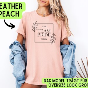 May include: A pink t-shirt with the text "2025 Team Bride Sophia" in a black square with floral accents. The model is wearing the shirt in an oversized style.