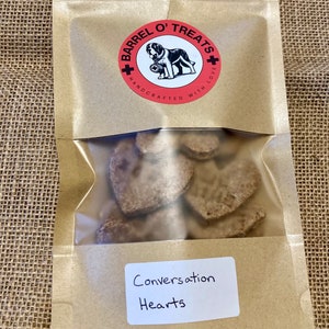 Pode incluir: Um saco de papel castanho com um logotipo vermelho e branco que diz "Barrel O' Treats Handcrafted With Love". O saco cont&eacute;m guloseimas para c&atilde;es em forma de cora&ccedil;&atilde;o e um r&oacute;tulo branco que diz "Conversation Hearts".