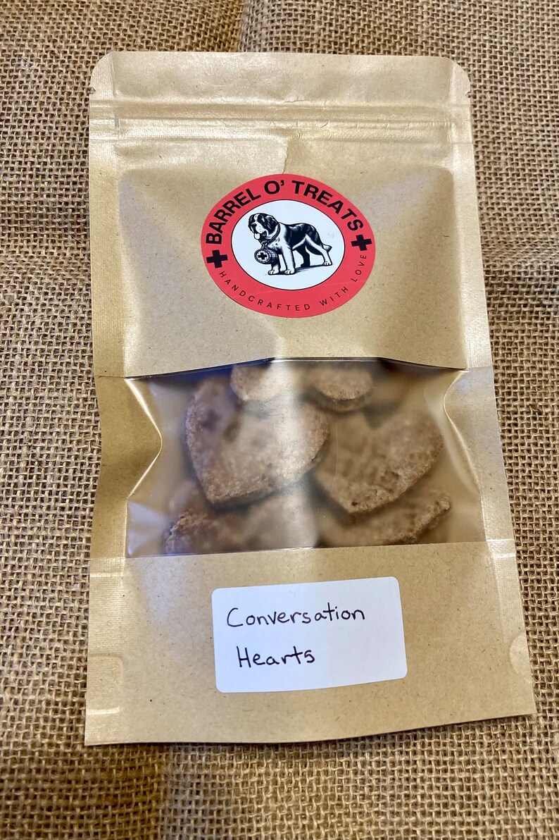 Pode incluir: Um saco de papel castanho com um logotipo vermelho e branco que diz "Barrel O' Treats Handcrafted With Love". O saco cont&eacute;m guloseimas para c&atilde;es em forma de cora&ccedil;&atilde;o e um r&oacute;tulo branco que diz "Conversation Hearts".