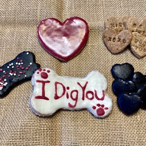 May include: Assortment of dog treats in the shape of hearts and bones. The treats are decorated with red, white and black icing and sprinkles. One bone-shaped treat has the words "I Dig You" written on it.